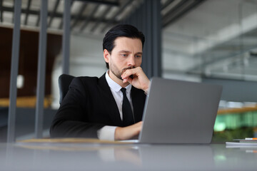 Wall Mural - A man in a suit is sitting at a desk with a laptop in front of him. He is deep in thought, possibly working on a project or dealing with a difficult problem. Concept of focus and concentration