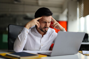 A man is sitting at a desk with a laptop open in front of him. He is looking at the screen with a puzzled expression on his face