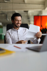 Wall Mural - A man is sitting at a desk with a white shirt and a piece of paper in his hand. He is smiling and he is happy