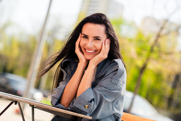 Sticker - Photo of charming positive woman wear grey trench hands cheekbones smiling enjoying walk sitting cafeteria outdoors urban city street