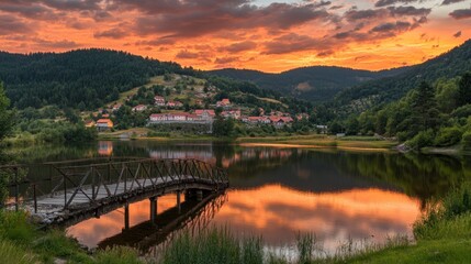 Wall Mural - Serene sunset over a tranquil lake with a wooden bridge and a picturesque village backdrop.