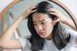 © miss irine - Young Asian woman examines her scalp in mirror, concerned about hair loss. Her hand is gently touching her thinning hairline. This image highlights common problem for many people balding, hair fall.