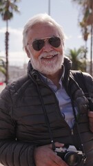 Wall Mural - Vertical. Grey hair senior man tourist with beard and sunglasses looking smiling at camera enjoying winter sunny vacation outdoor. Only older male people posing happy standing on pensioner holiday