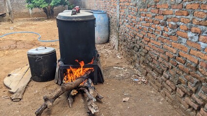 A large, black metal pot is placed on a makeshift stove made of bricks and wood. A fire is burning underneath the pot, producing flames and smoke. As well as a brick wall and a tree in the background.