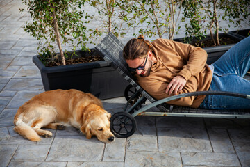 young attractive and sporty guy in casual clothes, lying on a sunbed outside in the home garden playing and having fun with his pet dog during a spring day at sunset