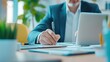 © JR-50 - A businessman writes notes while working on a laptop in a bright office with green plants and organized paperwork.