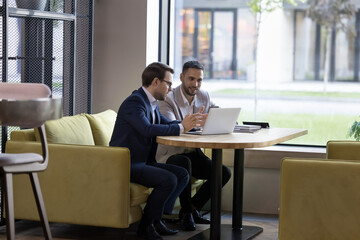 two engaged business partners talking at laptop in office cafe, watching online project presentation