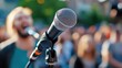 © Pinklife - A microphone stands ready on stage for an outdoor public speaking event, with a blurred crowd in the background on a bright sunny day, illustrating anticipation and focus.