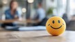 © Pinklife - A bright yellow stress ball with a smiling face sits on an office desk, promoting positivity and stress relief among employees, in a modern work environment background.