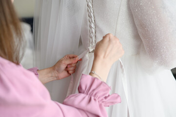 Morning preparation of bride. Bridesmaid helps bride to fasten, lace up white wedding dress. Close-up of girl hand tying bow on wedding dress