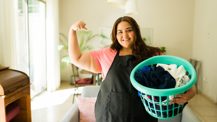 Strong woman wearing an apron smiling and showing her muscle while carrying a laundry basket full of clothes