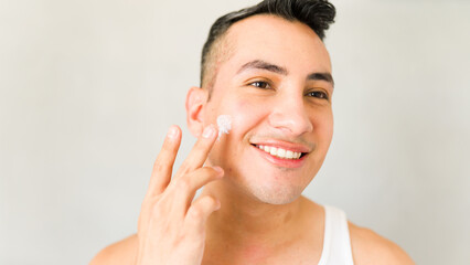 Young man promotes a healthy skincare routine for men by smiling while applying face moisturizer