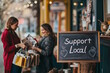 © MarGa - Two women shopping with 'Support Local' sign outside store, concept Small Business Saturday