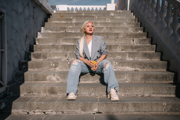 Wall Mural - A woman in a suit and jeans sits on a set of stairs. She is holding a cup and she is smiling.