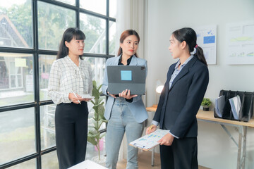 Three asian businesswomen in a bright office, discussing work by a window with a laptop and documents, showcasing teamwork and communication for corporate success