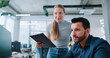 © ihorvsn - Camera view of people working together in office. Handsome man sitting at computer while girl standing next to him with clipboard in her hands. Caucasian coworkers making working task together.