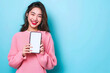 © Humans on Color BG - Full body young smiling happy woman of Asian ethnicity 20s in pink sweater stand near big mobile cell phone with blank screen workspace area chatting isolated on pastel plain light blue background.