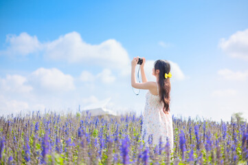 A young woman in a floral dress takes photos in a lavender field under a bright blue sky. The serene scene captures the joy of photography, nature, and sunny weather
