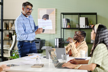 Aufkleber - Middle-aged man with glasses showing charts on printed paper during business meeting while colleagues attentively listening and discussing in modern office space