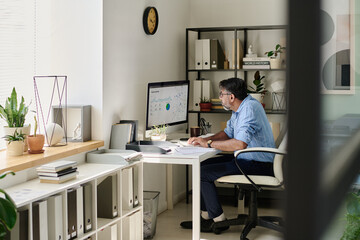 Poster - Senior man analyzing data on computer in modern office setting featuring shelves, plants, and decor items. Mid-century modern style felt throughout the room harmoniously