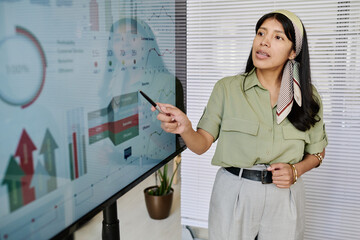 Poster - Woman wearing headscarf presenting data on large screen in office setting Holding pointer, she is discussing financial and analytical growth charts indoors, possibly during business meeting