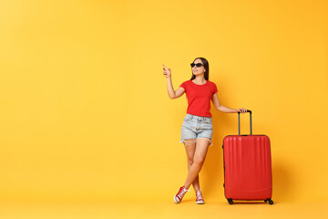 Canvas Print - Happy young woman in sunglasses with suitcase pointing at something on orange background