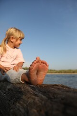 Canvas Print - Cute little girl sitting on tree trunk near river, selective focus. Child enjoying beautiful nature