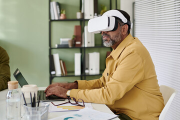 Poster - Senior man engrossed in virtual reality experience while seated at desk in modern office. Blurred background with shelves and blinds in view