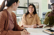 © pressmaster - Smiling woman wearing glasses engaging colleagues at meeting table in modern office with large windows. Multiracial team collaborating in workspace filled with natural light and technology