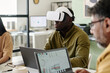 © pressmaster - Man immersed in virtual reality using VR headset while sitting in contemporary office space alongside two colleagues collaborating on digital devices