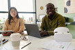 © pressmaster - Two colleagues, one Black person and one Caucasian, smiling and collaborating on project with laptop and documents on table, enjoying bright and open workspace