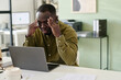 © pressmaster - Businessman holding his head while working on laptop, showing stress and frustration in modern office environment with documents on desk