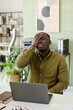 © pressmaster - African American man feeling stressed while working in modern office with laptop on desk. Background includes shelves, plant, and other office supplies creating professional environment