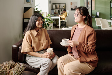 Poster - Two women having coffee and conversation in office lounge area, exchanging smiles and ideas while sitting on comfortable chairs in a well-lit office environment