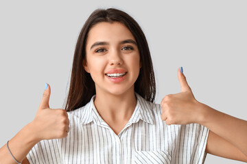 Wall Mural - Smiling young woman with healthy teeth showing thumbs-up on light background, closeup