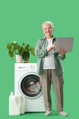 Wall Mural - Happy senior woman with laptop doing laundry on green background