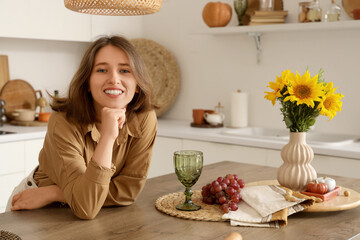 Poster - Pretty young woman leaning on table with autumn flowers, grapes and pumpkins in kitchen