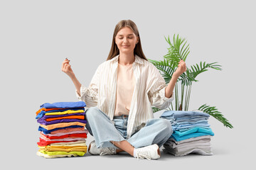 Young woman with stacks of clothes and palm tree meditating on light background