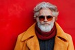 © Nataliia_Trushchenko - Man in a yellow coat and red scarf stands in front of a red wall. He is wearing sunglasses and has a beard. Concept of old men's health, self care, fashion and beauty, healthy lifestyle.