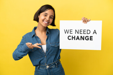 Young mixed race woman isolated on yellow background holding a placard with text We Need a Change and  pointing it