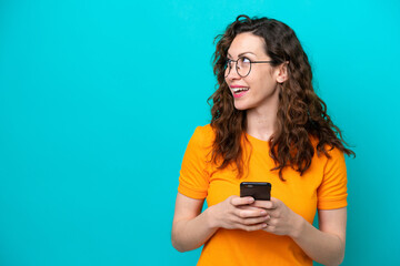 Wall Mural - Young caucasian woman isolated on blue background using mobile phone and looking up