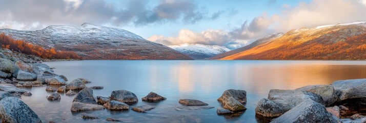 Wall Mural -  A large body of water, encircled by rocks, lies beneath snow-capped mountains under a cloudy yet blue sky, dotted with a few clouds