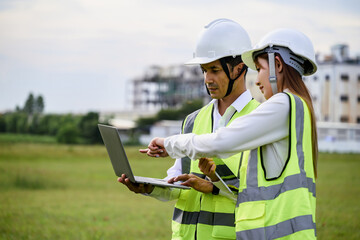 Two engineers wearing safety gear discuss project details while referencing a laptop. They stand outdoors with a blurred industrial background.