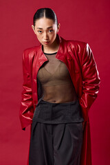 Wall Mural - Asian woman in red leather jacket and black pants posing against red background with eyes closed and hands on hips