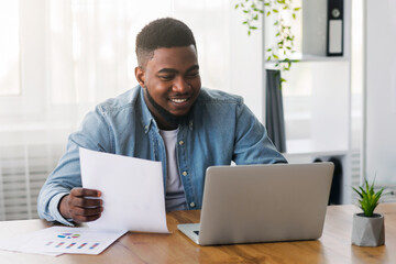 Poster - Cheerful african american businessman working with laptop and papers in office, preparing financial report. Copy space