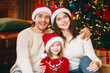 © Prostock-studio - Affectionate young family in Santa hats sitting by Christmas tree together, hugging and smiling at camera