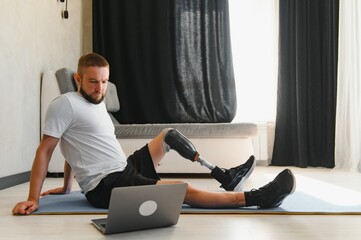 Sporty fit young man amputee with prosthetic leg disability prosthesis doing training exercises on mat at home using laptop. Inclusive online sport classes for people with disabilities concept