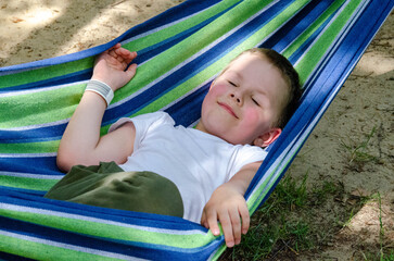 Cute little blond caucasian boy relaxing and having fun in multicolored hammock in backyard or outdoor playground. Summer active leisure for kids. Child swinging on hammock. Activities for children