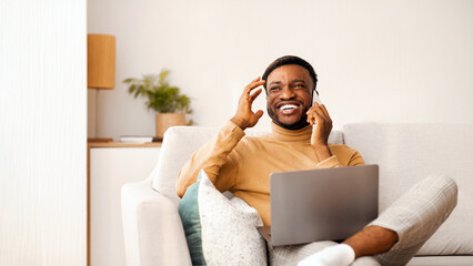 Canvas Print - Positive Afro Man Using Laptop Talking On Mobile Phone And Laughing Sitting On Sofa Indoor. Selective Focus, Copy Space