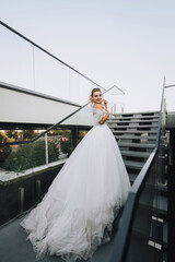 Beautiful young curly smiling blonde bride in white long dress standing on steps of stairs in city outdoors. Wedding photography.